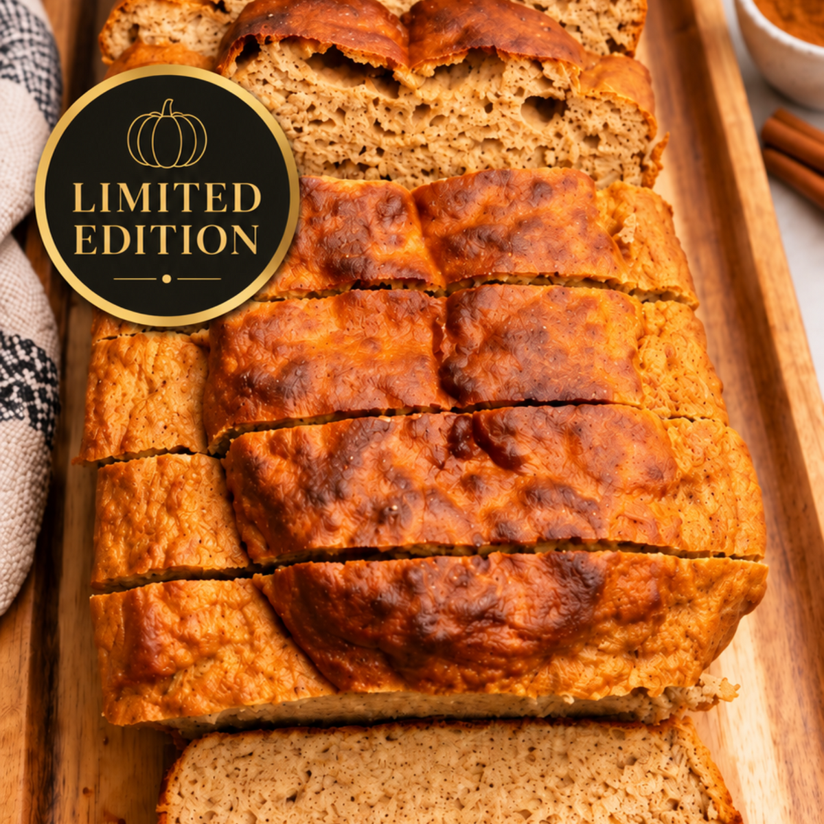 A loaf of sliced pumpkin bread on a wooden serving board, with a round black and gold 'LIMITED EDITION' badge overlaid on the left side. A small bowl of cinnamon and a striped cloth are partially visible in the background.