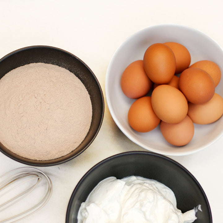 Bowls of flour, eggs, and cream on a white surface with a whisk.